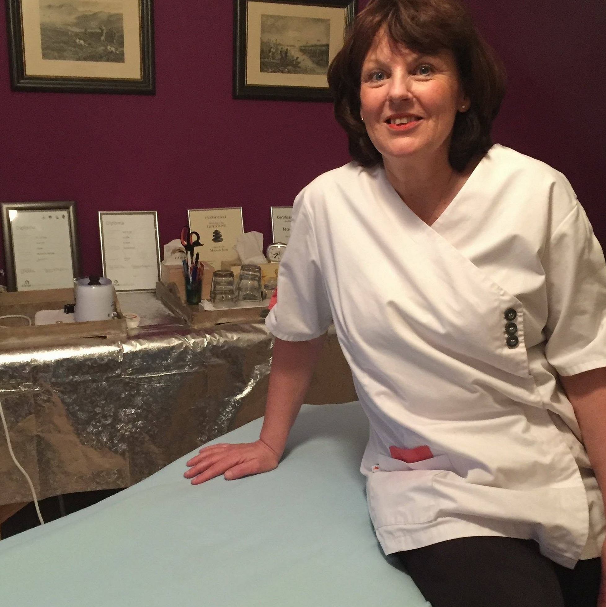 Marja de Jong in een witte tuniek, zittend op de massagetafel in haar praktijk
